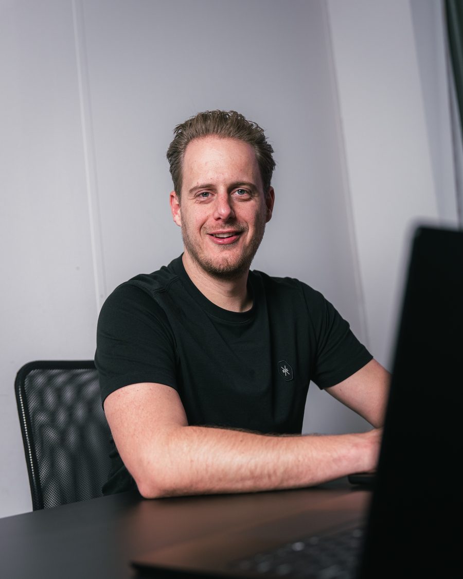 Maurits den Dunnen zit lachend achter zijn laptop en kijkt in de camera